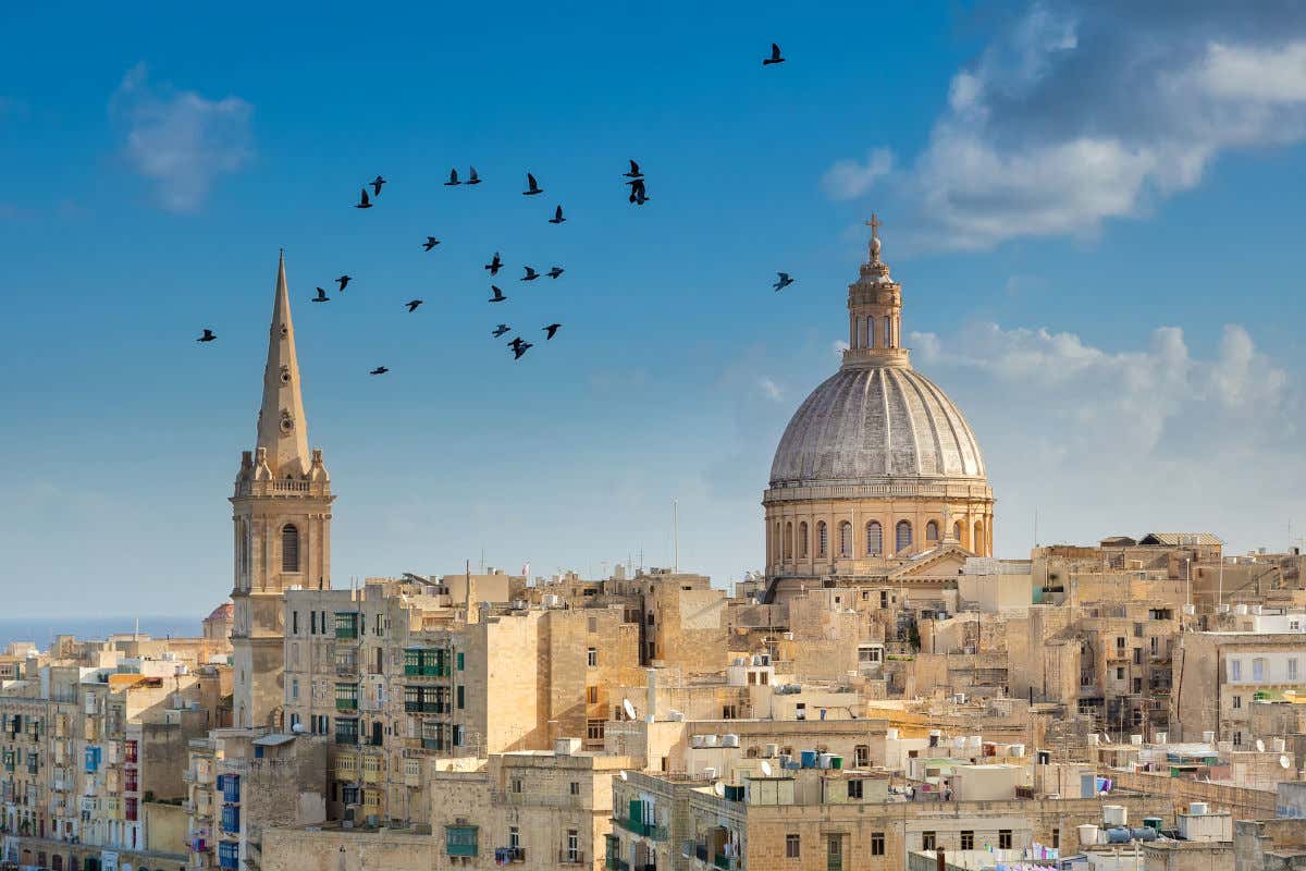 Panorámica de la ciudad de La Valeta con varios edificios con la cúpula de una catedral y una torre en primer plano y varios pájaros sobrevolando la escena