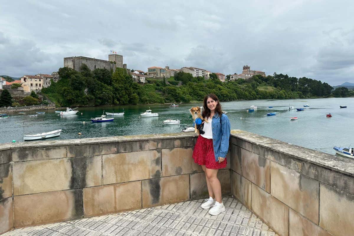 Rocío Molla sosteniendo un pequeño perro en brazos en un mirador de San Vicente de la Barquera con vistas al mar a un castillo