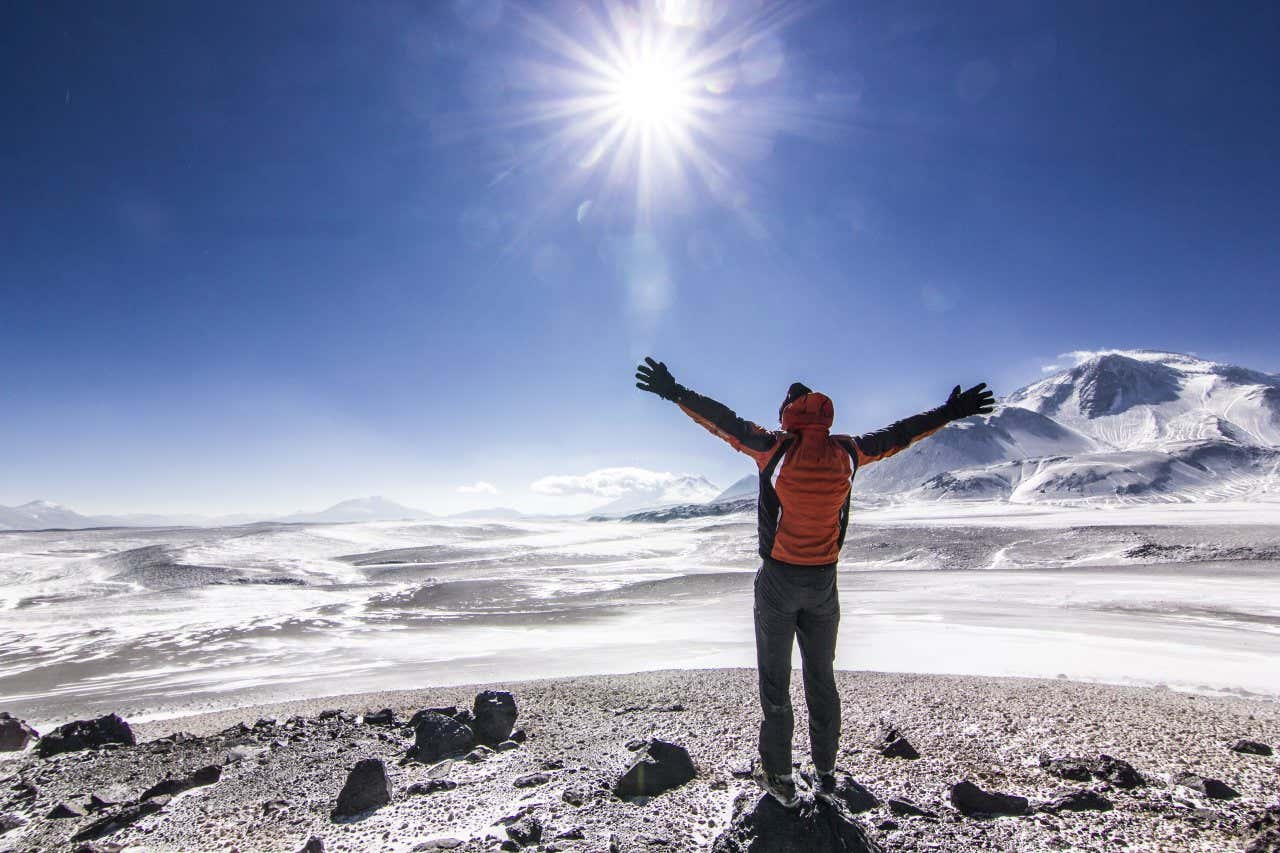 Um homem com os braços abertos contemplando a paisagem nevada do vulcão Ojos de Salado em um dia ensolarado