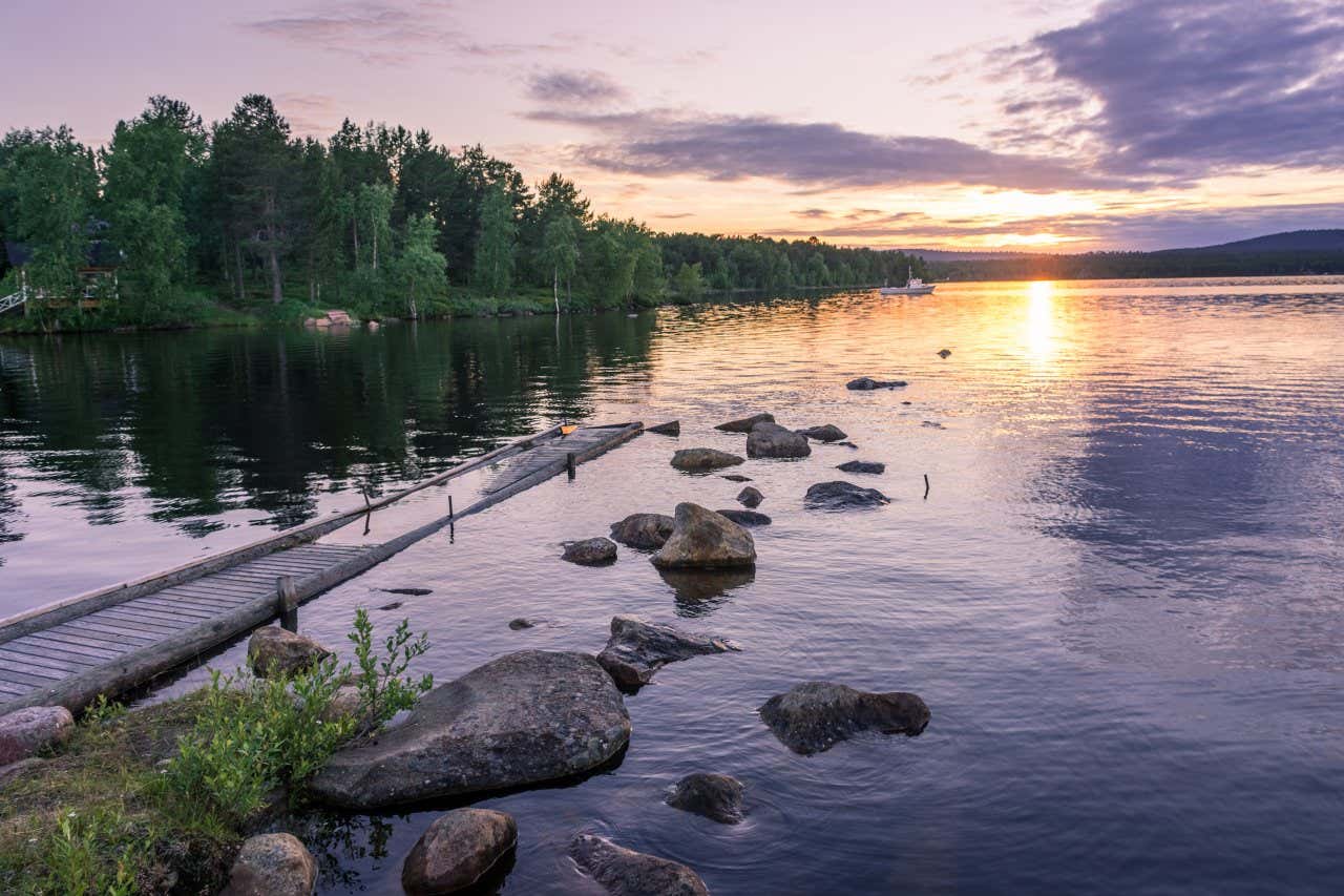 El lago Inari en Finladia con un embarcadero parcialmente hundido y un barco navegando junto a una orilla repleta de árboles