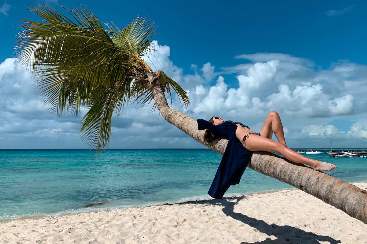 Una mujer en bikini tumbada sobre una palmera en una playa de arena blanca con aguas cristalinas