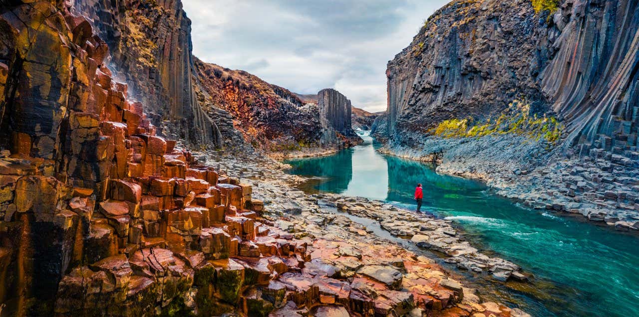 Escursionista con giubbotto rosso vicino a un fiume cristallino tra un canyon dai colori rossi e grigi sotto un cielo nuvoloso in Islanda