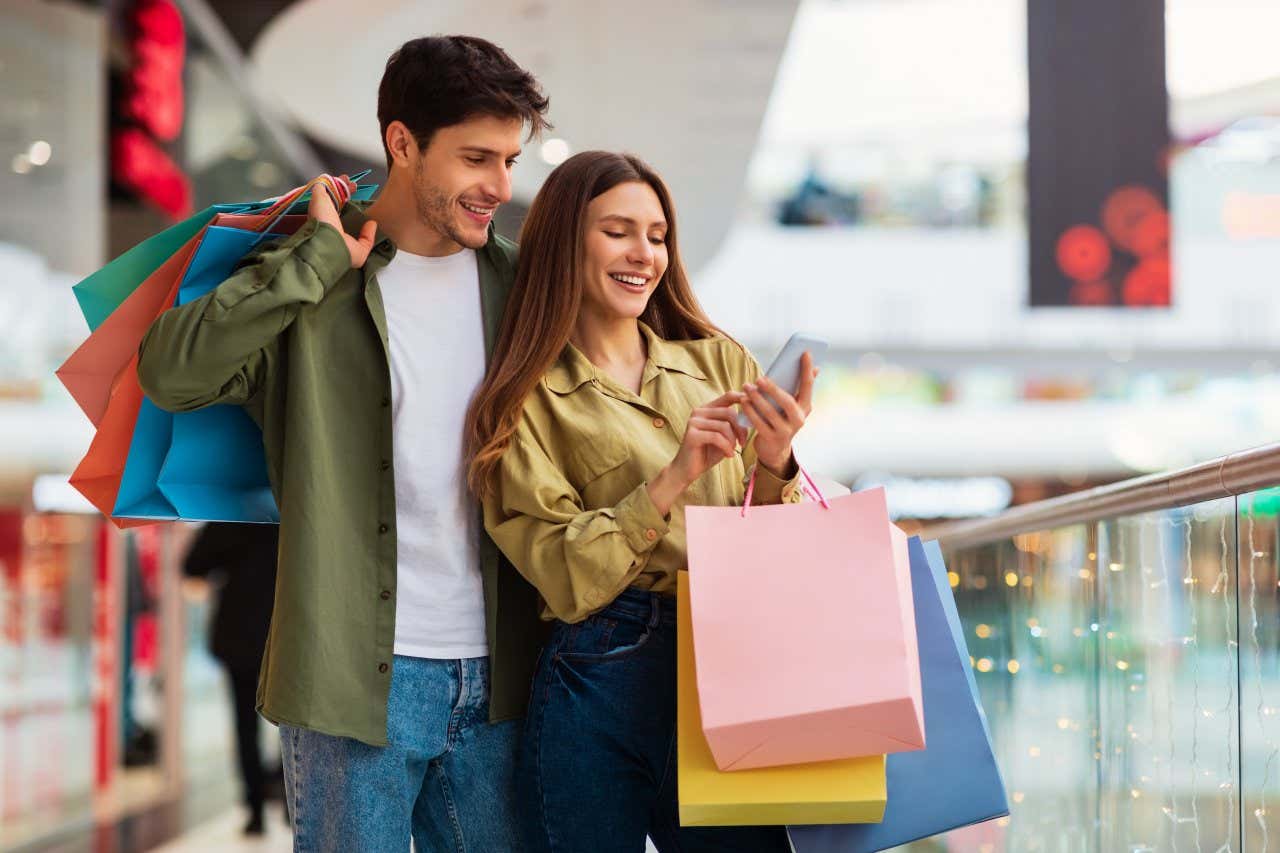 Couple faisant du shopping en utilisant des téléphones portables et en tenant des sacs colorés dans un centre commercial