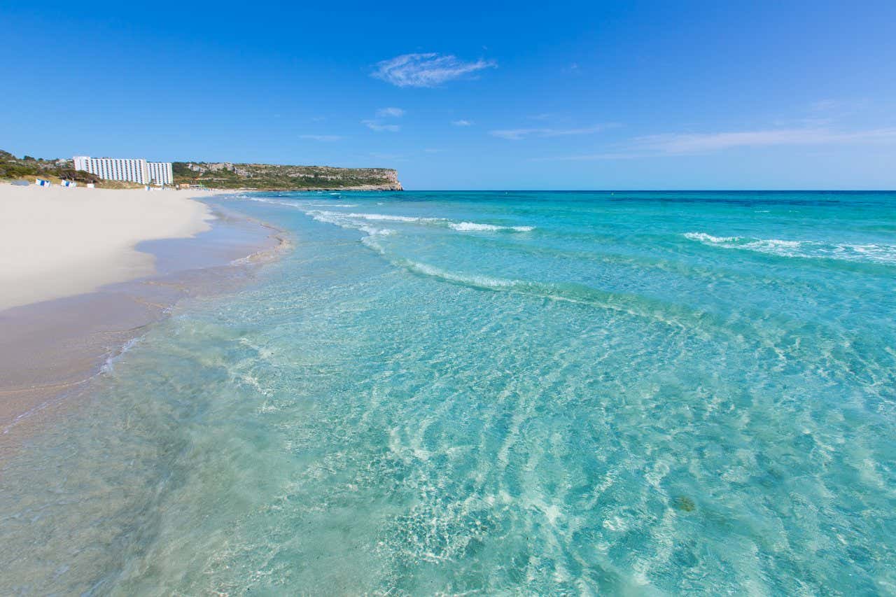 Son Bou Beach with waves coming in on the white sand, and a clear sky in the background.