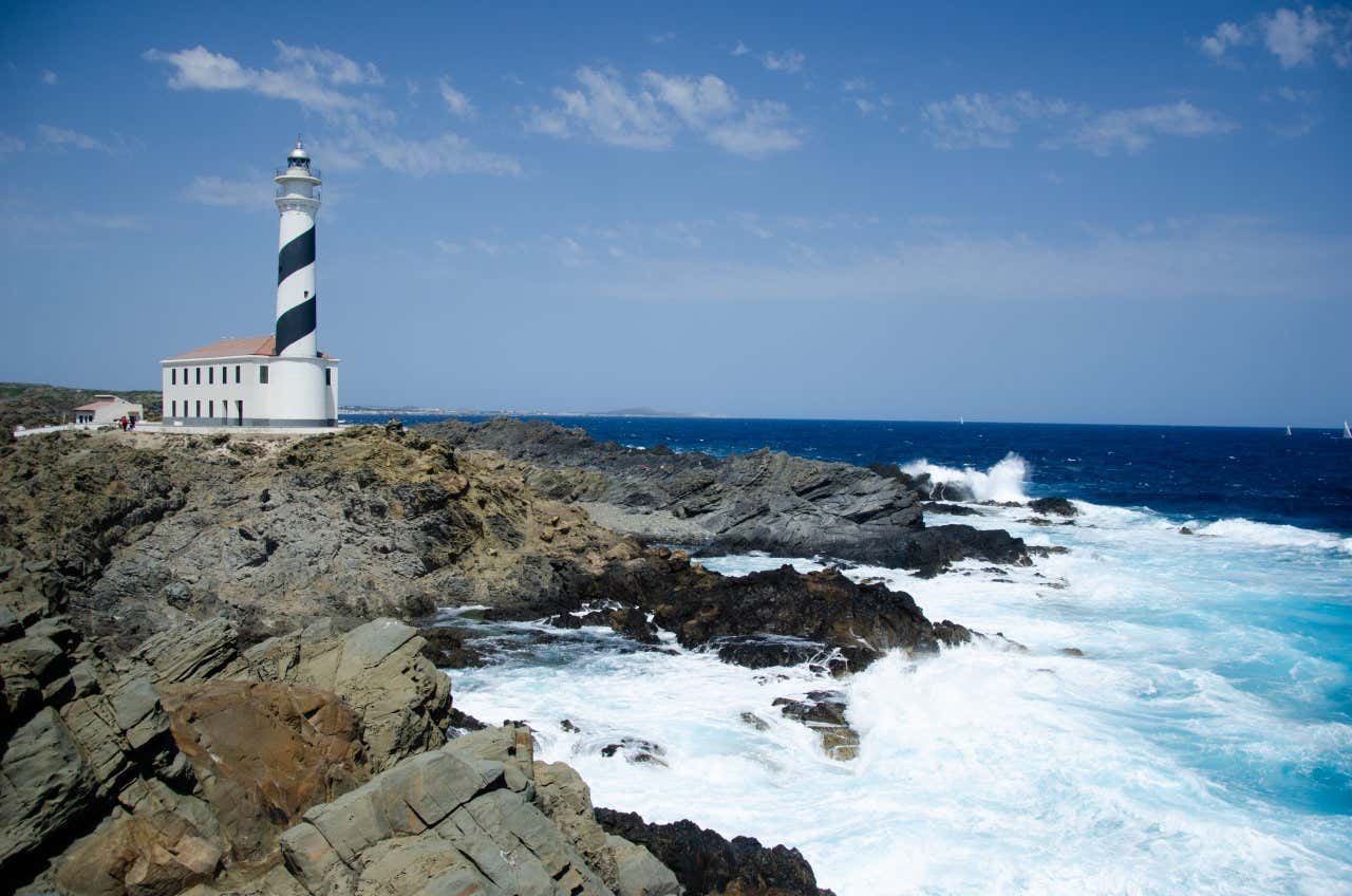 Favàritx Lighthouse, with distinctive black band around it looking over waves thrashing onto the Menorcan coastline,