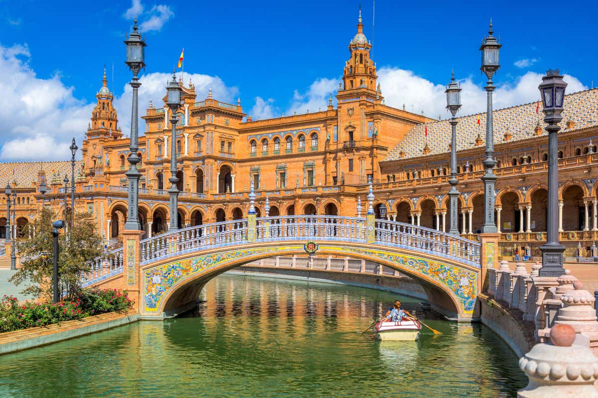 Una barca con dos turistas navegando junto a uno de los puentes de la Plaza de España de Sevilla en un día prácticamente despejado