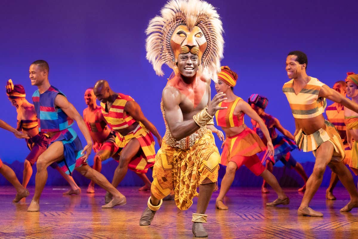 Un acteur portant un masque de lion et de nombreux danseurs en costumes ethniques sur une scène de Broadway lors d'une comédie musicale new-yorkaise