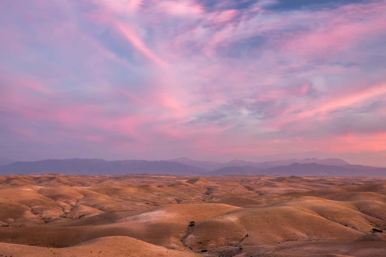 The Agafay Desert in Morroco with a pink and purple sky and a mountain range in the background.
