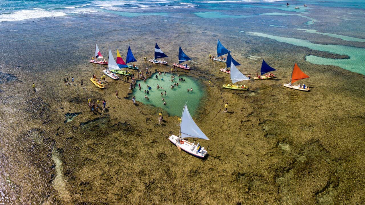 Vista aerea delle piscine naturali di Porto de Galinhas con persone che nuotano e diverse imbarcazioni a vela colorate