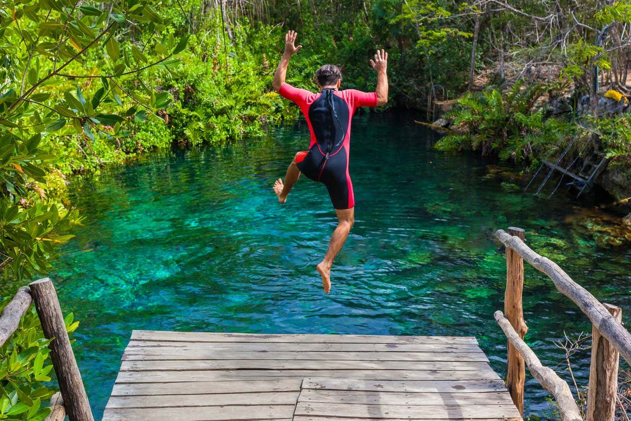 Um homem vestido com neoprene pulando da ponte de mandeira ao cenote 