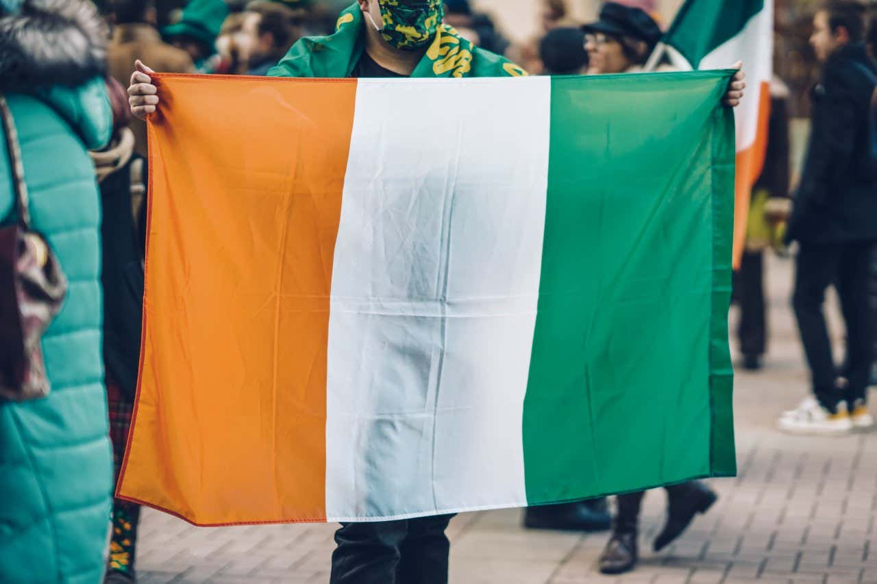 Uma pessoa usando uma máscara verde, preta e amarela e segurando a bandeira da Irlanda.