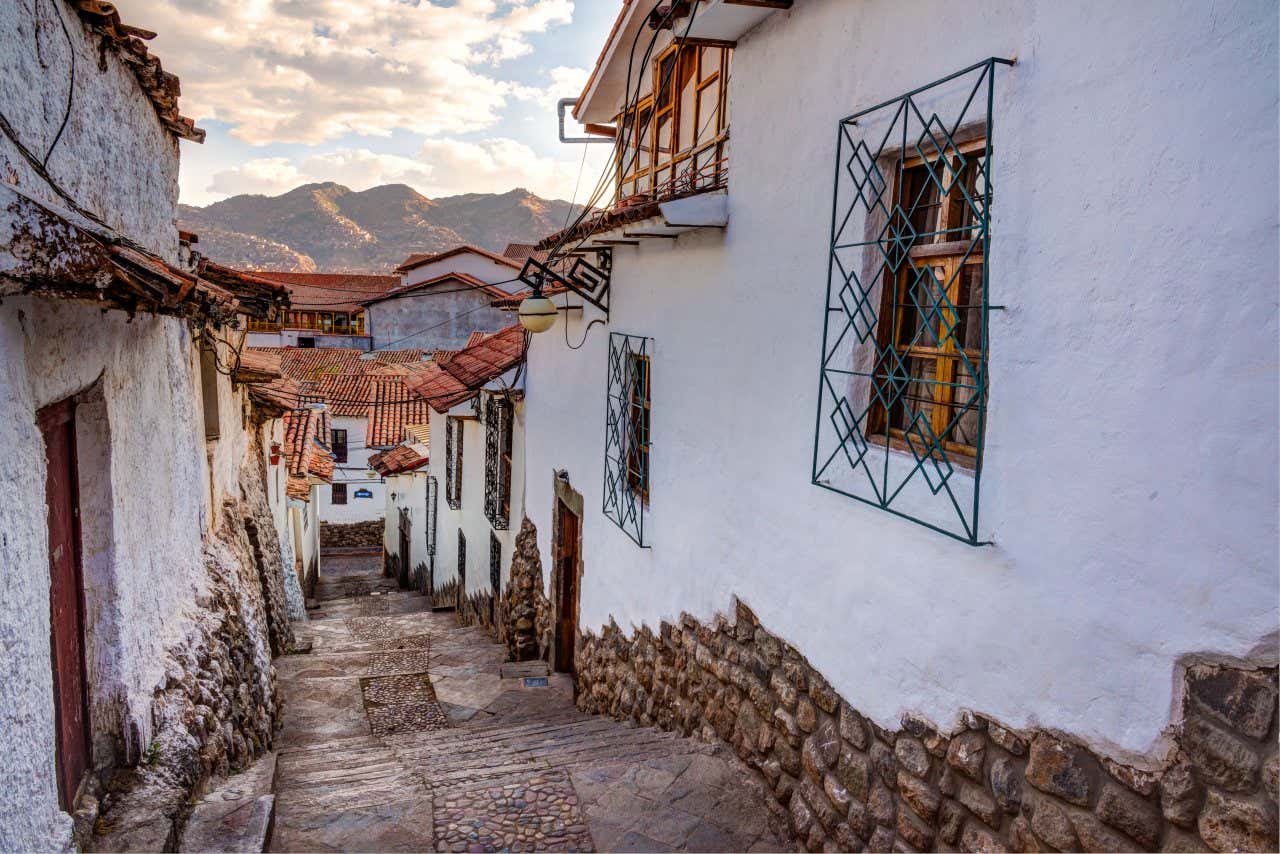 A narrow street with steps as seen from the top, with a cloudy sky and a mountain range visible in the distance.