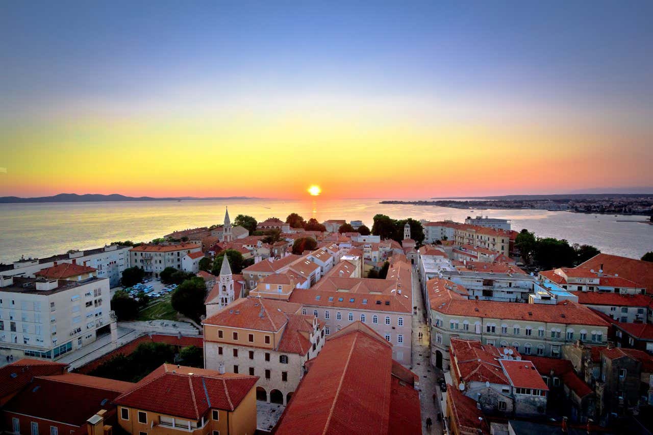 Uma panorâmica aérea de Zadar, com o sol se pondo ao fundo, em um céu azul e laranja