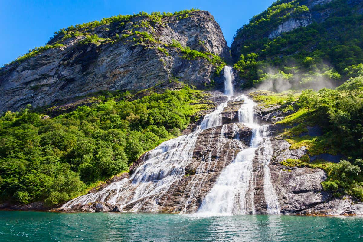 A white waterfall falling down grey cliffs between green forests under a clear blue sky