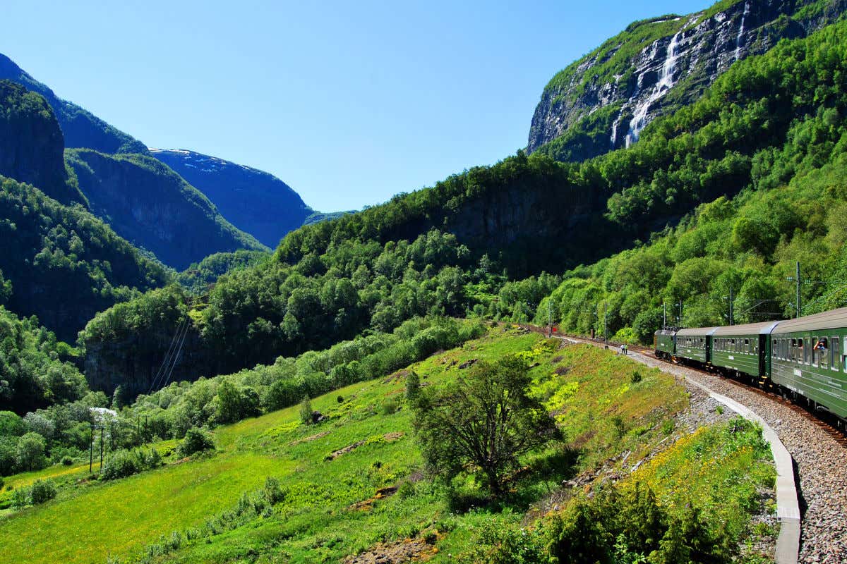 A dark green train passing through green forests and mountains with a waterfall on the right
