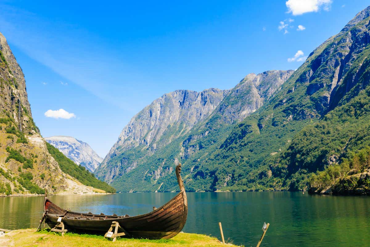 A small wooden viking boat by the shore of a fjord, surrounded by steep grey mountains covered in green forests