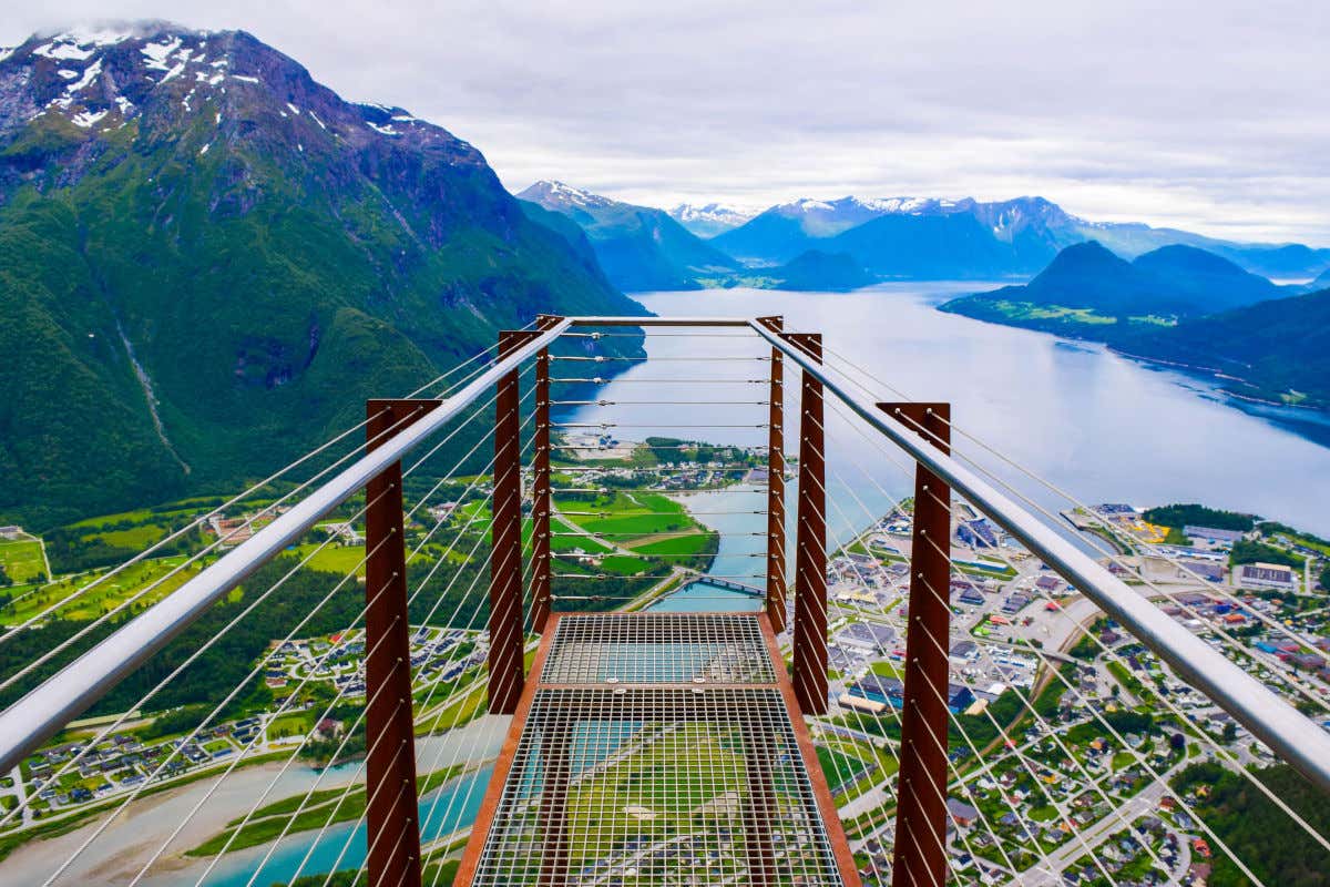 A narrow metal viewpoint over a town on the shore of a fjord, with mountains in the background