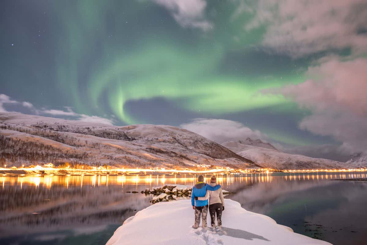 A couple in warm clothes hug stood in a patch of snow on the shore of a lake under green lights in the night sky