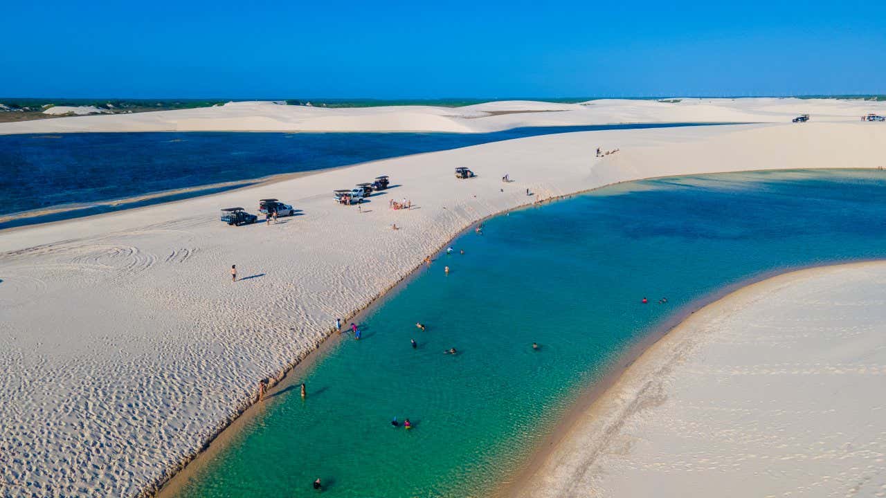 Des touristes se baignent dans une lagune turquoise et d'autres profitent du sable blanc des dunes avec quelques voitures 4x4 à proximité