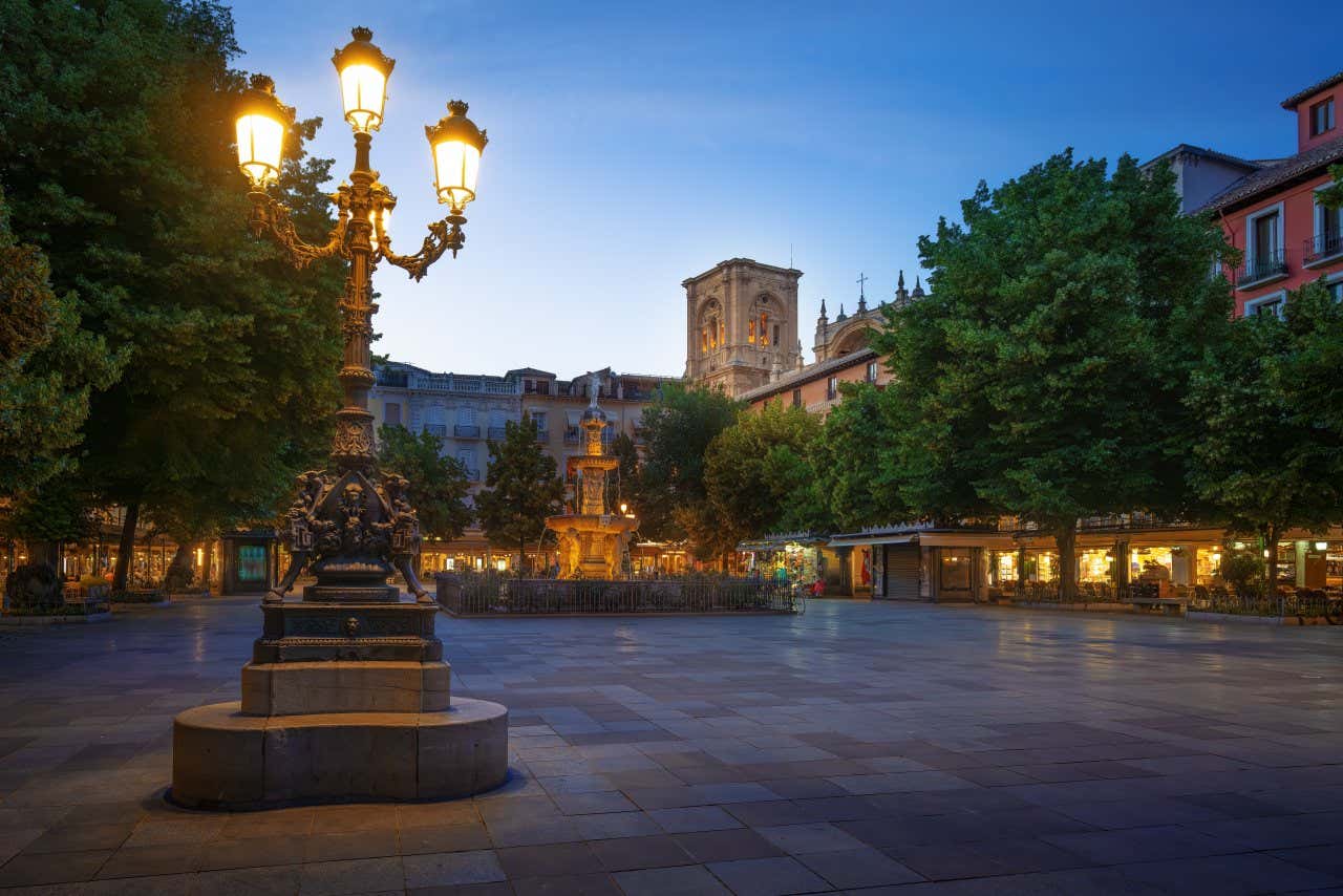 A shot of the Plaza de Bib-Rambla at night, with a lit lampost in the foreground and a fountain and sky in the background.