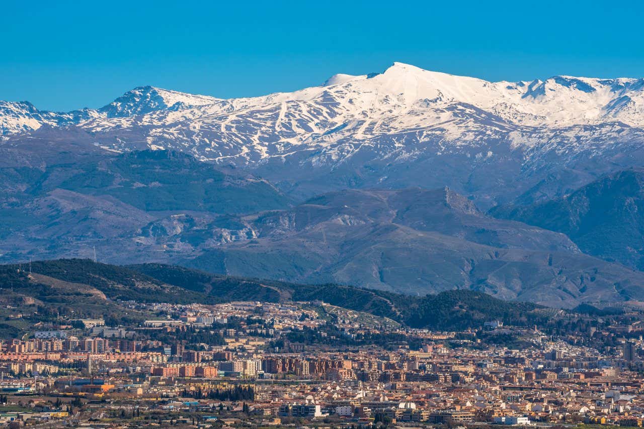 A shot of the snow-capped Sierra Nevada mountains as seen from afar, with a city in the foreground and a clear blue sky in the background.