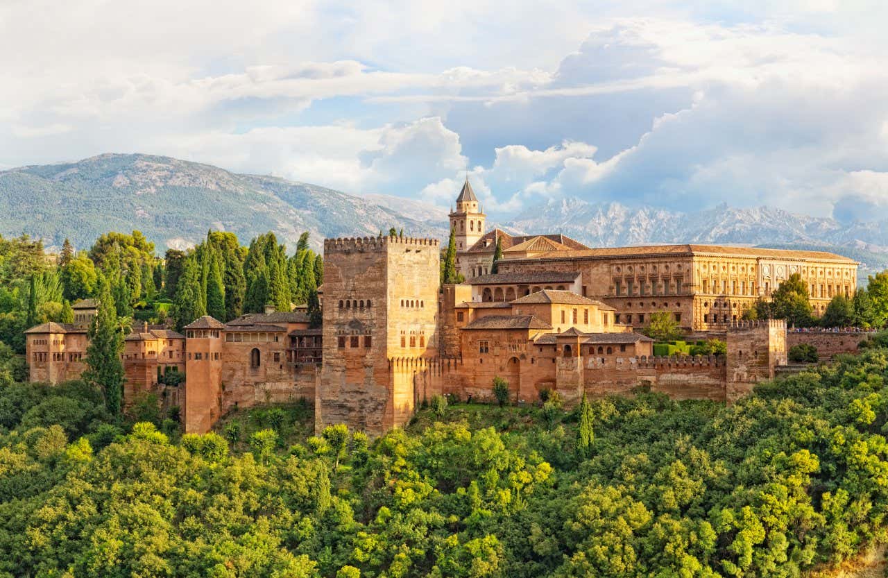 An aerial shot of the Alhambra complex, with vegetation seen below and mountains and a cloudy sky in the background.