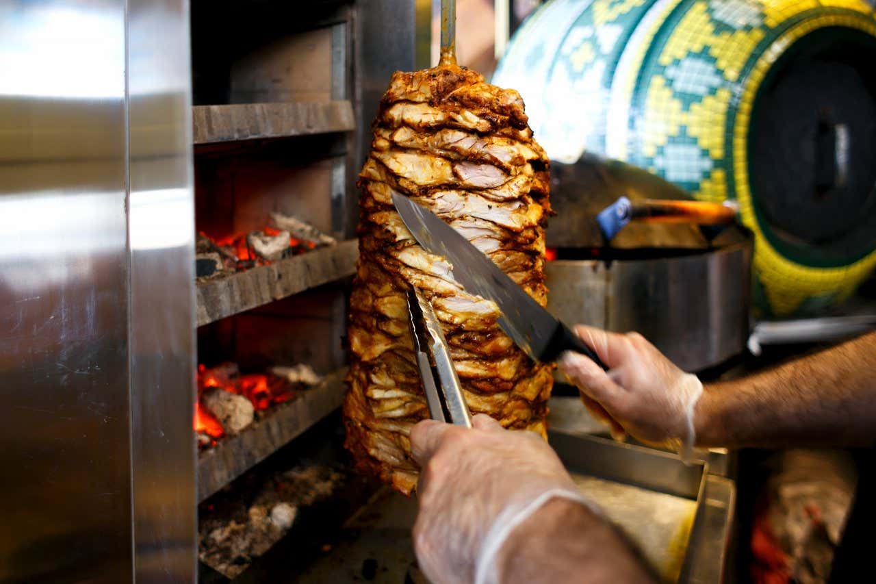 Um chef usando luvas de plástico e cortando a carne que está em um espeto vertical giratório.