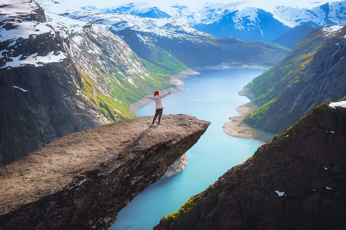 A woman with her arms stretched wide, stood on a pertruding rock over a large Norwegian Fjord with views of snowy mountains