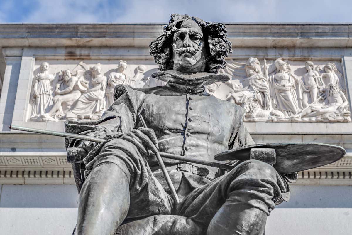 Escultura de bronce de Velázquez sosteniendo un pincel y una paleta de pinturas frente a un relieve escultórico de la fachada del Museo del Prado