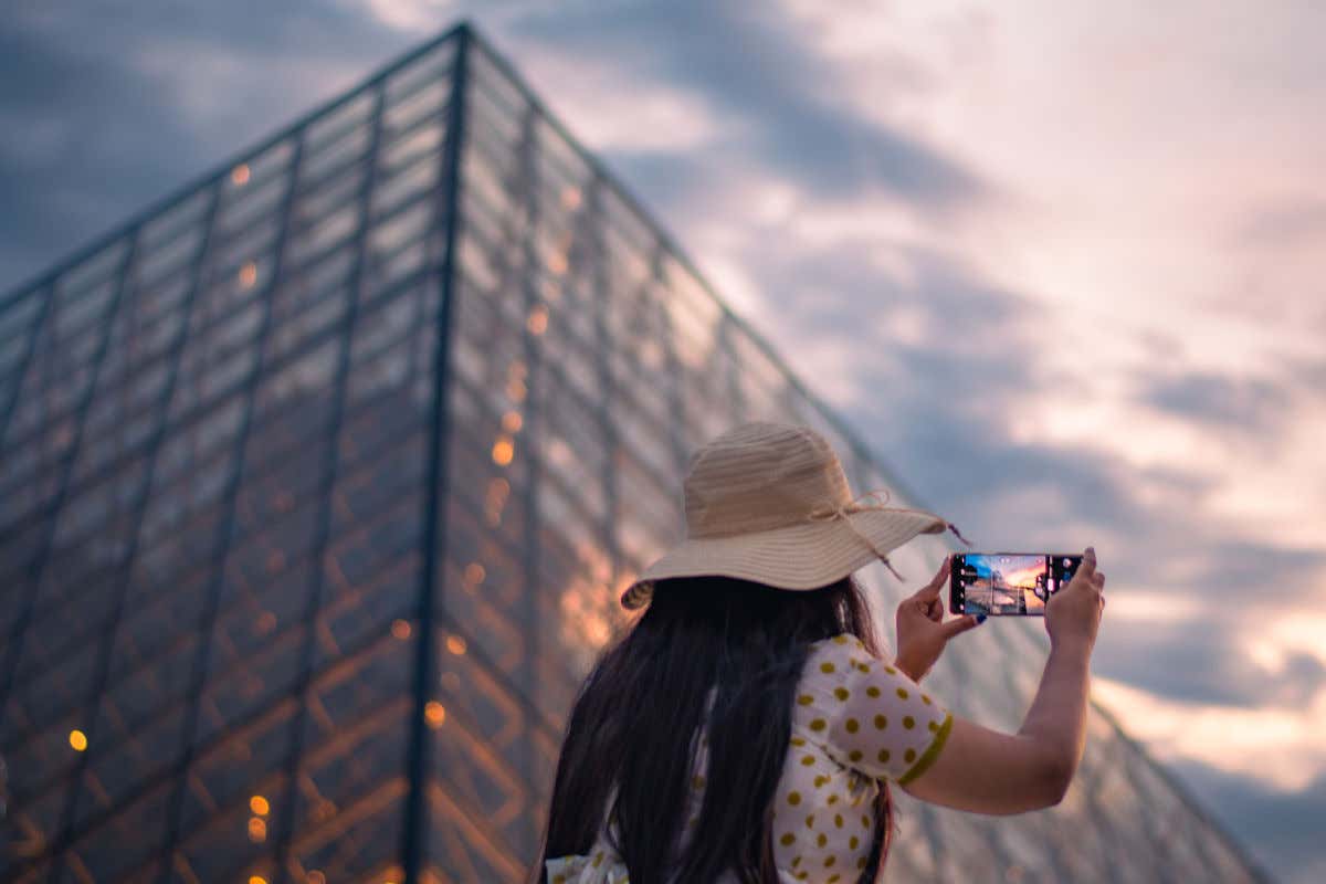 Una mujer tomando una fotografía desde el móvil de la pirámide de cristal del museo Louvre de París al atardecer