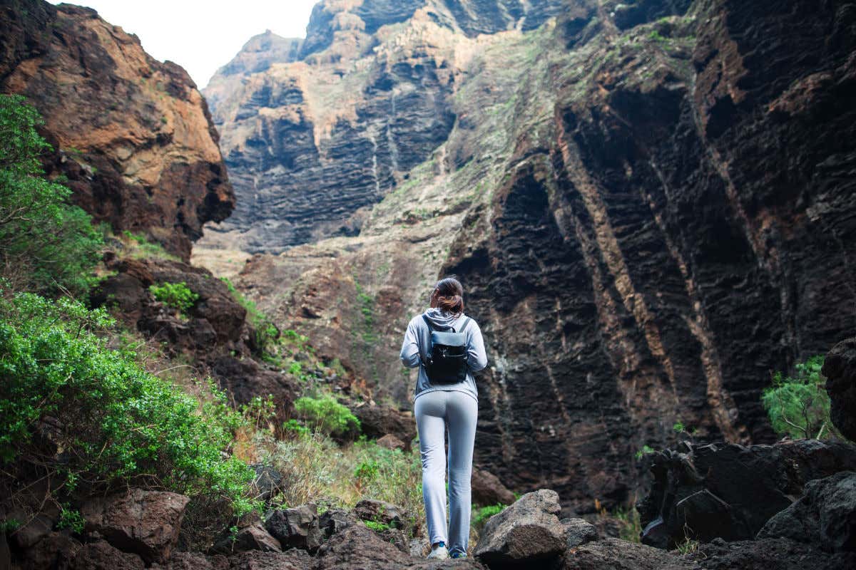 Una mujer con ropa deportiva de espaldas a la cámara participando en una ruta de senderismo que atraviesa un alto barranco de escasa vegetación