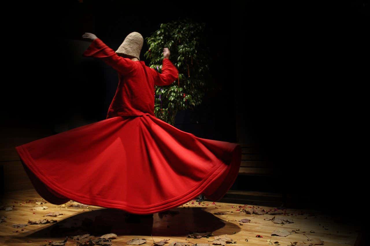 Danseur avec une longue robe rouge et un chapeau marron en train de danser dans une salle sombre