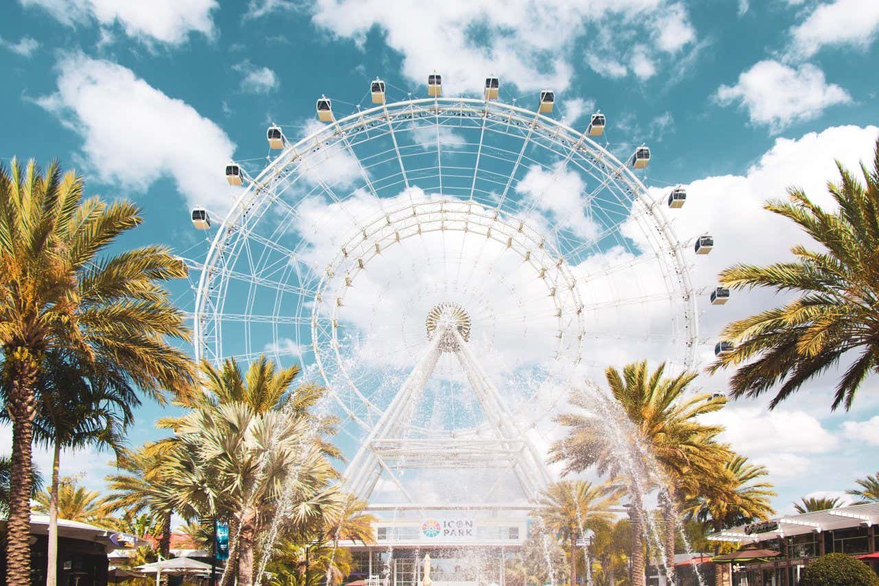 Grande roue avec une fontaine devant et des palmiers sous un ciel peu nuageux