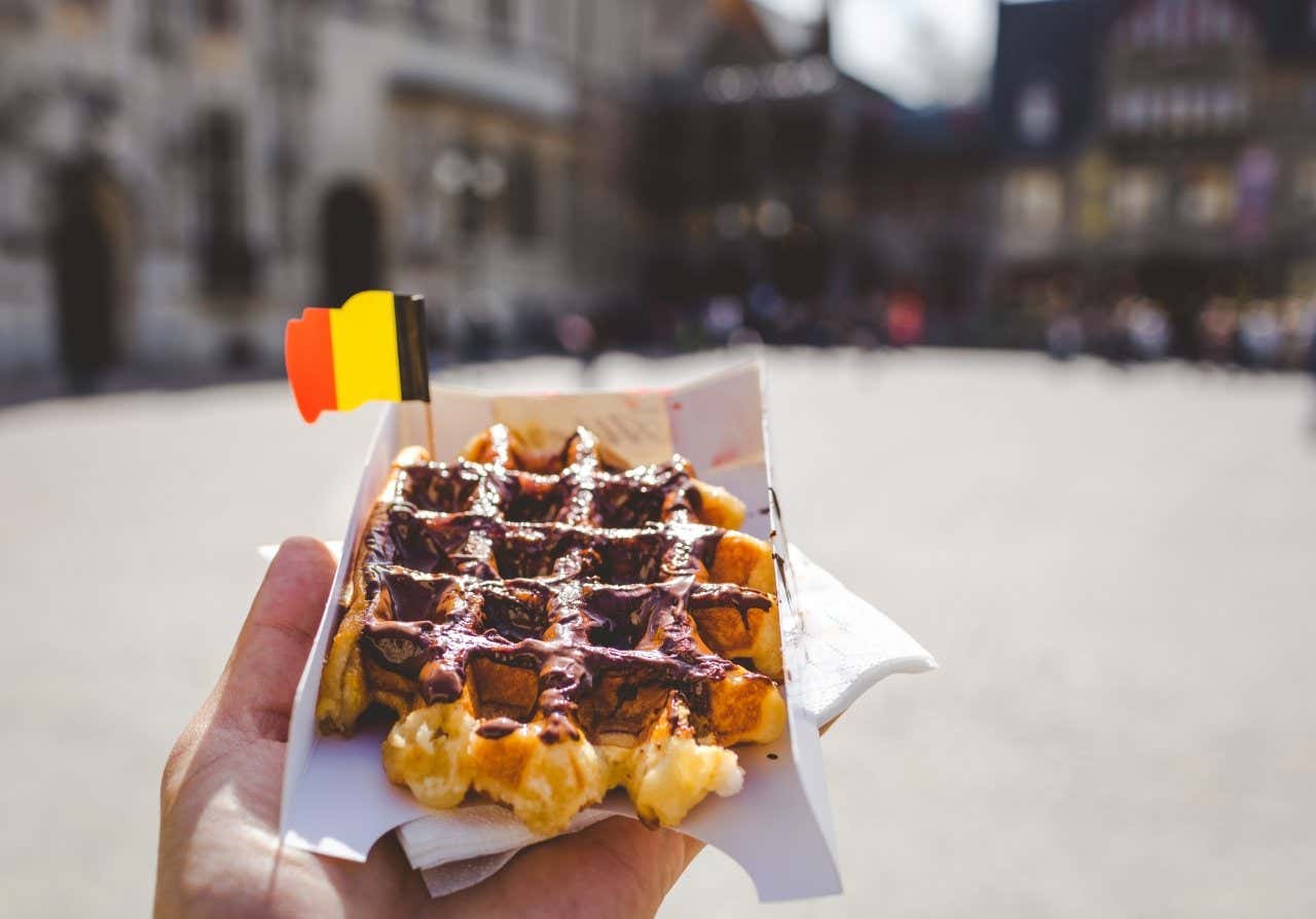 A hand holding a waffle covered in chocolate on a white carton with the Belgian flat in it in Bruges