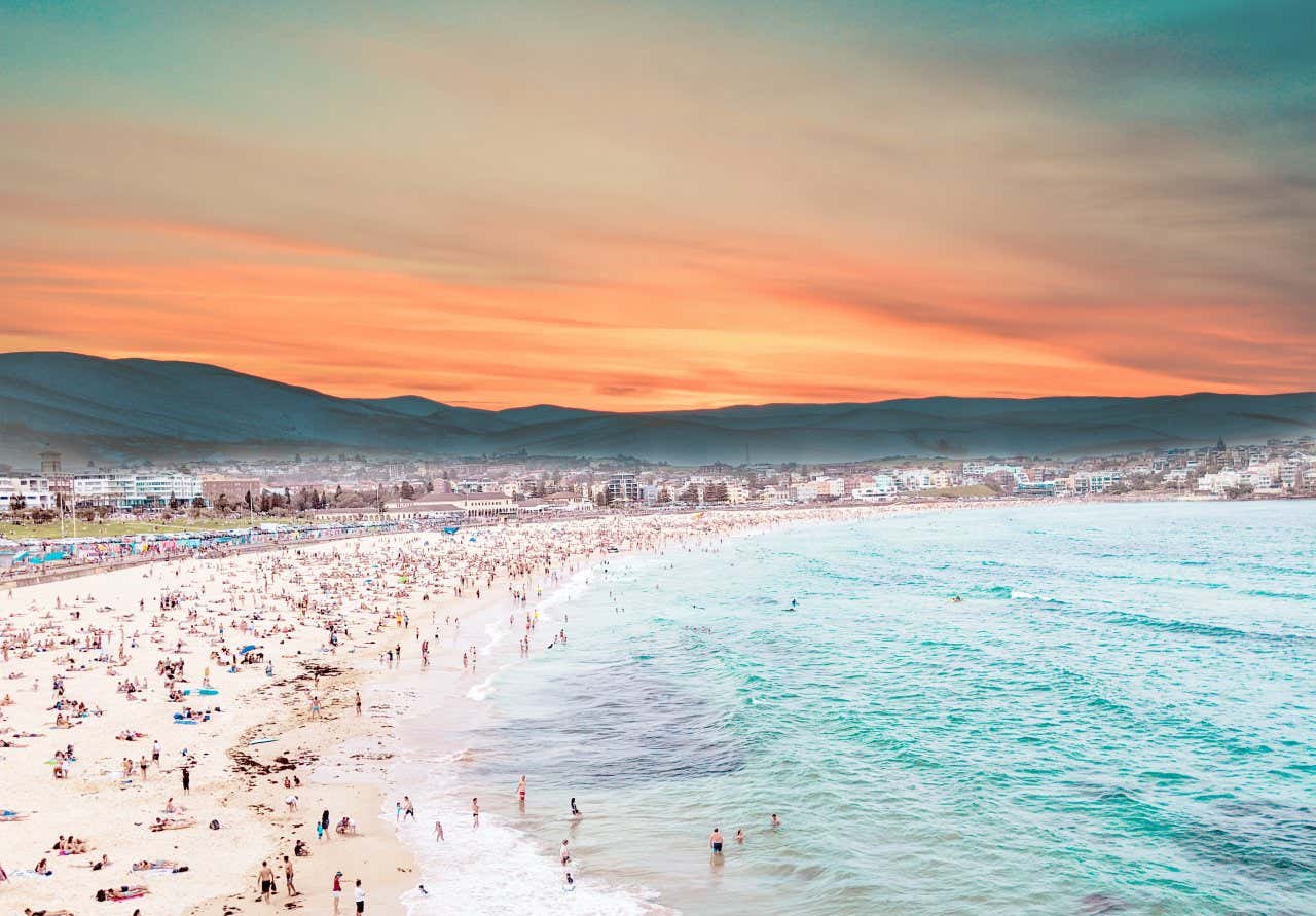 An aerial view of Bondi Beach, with a high concentration of beach-goers on the sand, and some in the water. An orange sky above mountains in the background.