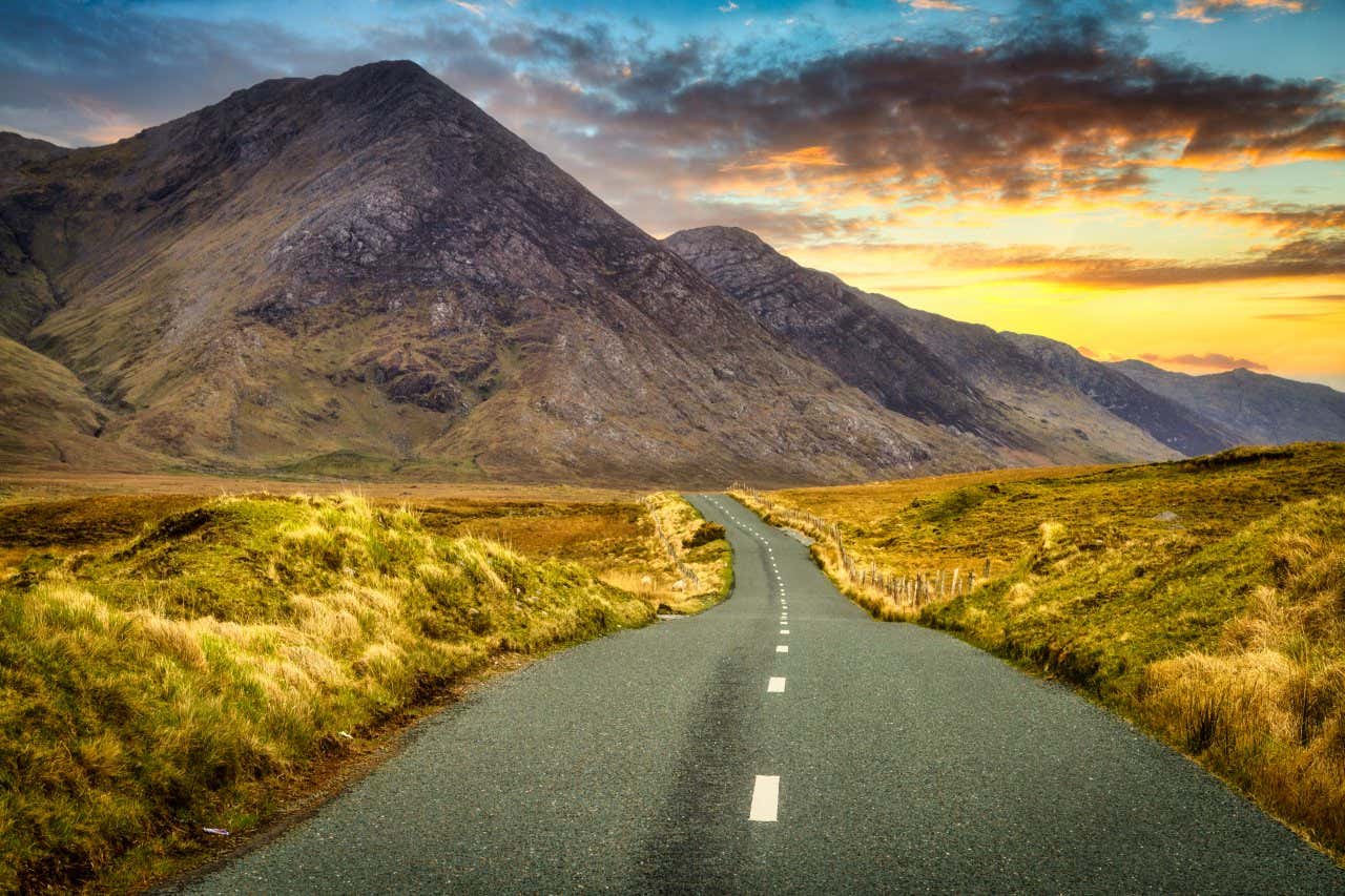 A narrow, winding, two-way road in Connemara, Ireland, with a cloudy yellow and blue sky in the background in view.