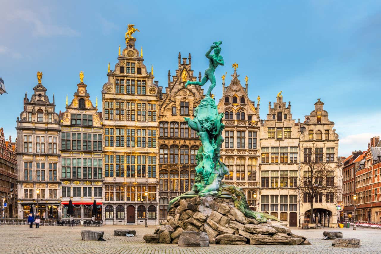 A copper statue on rocks in the centre of a cobbled square in Bruges, with tall typical Flemish houses behind