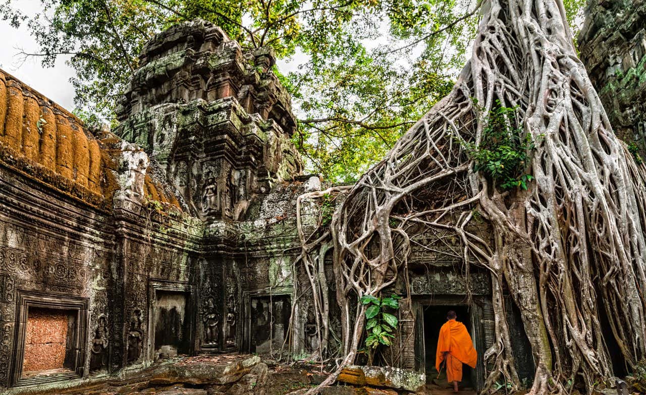 Un monaco vestito di arancione nel tempio di Angkor ricoperto da radici di alberi e circondato da antiche mura