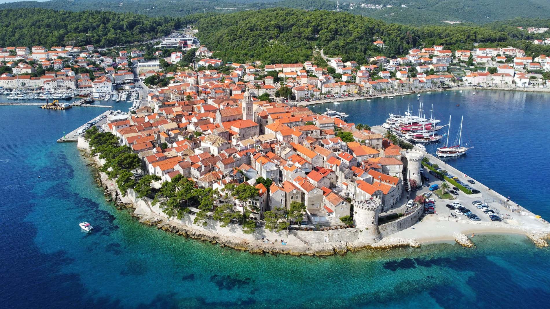 Vista aerea di Curzola con barche nel porticciolo e mare cristallino, con rigogliosa vegetazione sullo sfondo