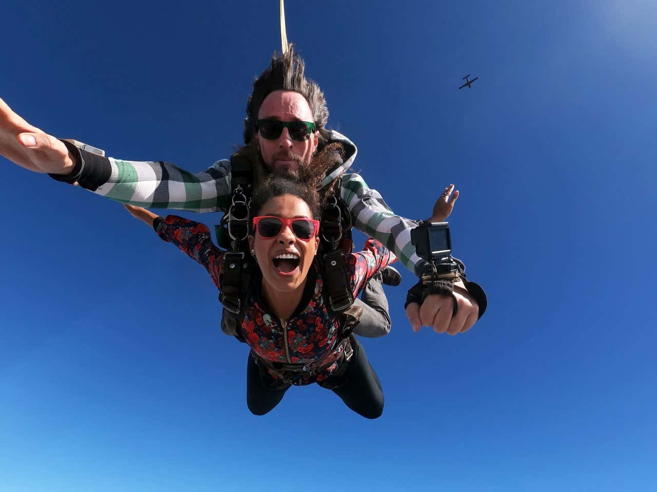 Two people skydiving as seen from below, with a plane in view in the clear blue sky above.