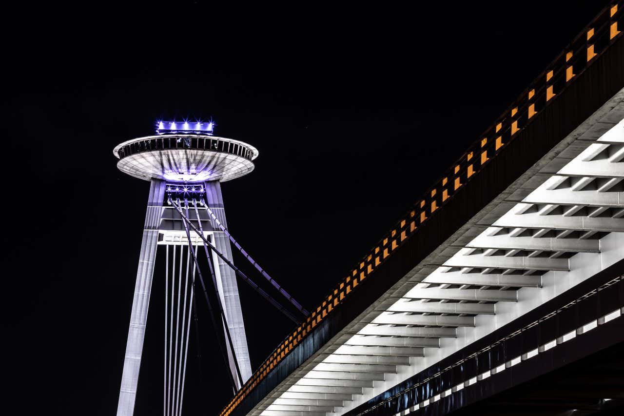 Uma foto da Torre UFO, iluminada em roxo, em Bratislava, à noite, tirada de baixo da Ponte Nova.