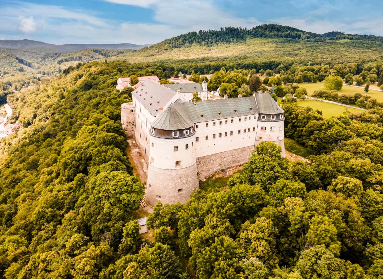 Uma foto aérea do Castelo de Cerveny Kamen, cercado por árvores, com um céu nublado visível na parte superior da foto, ao fundo.