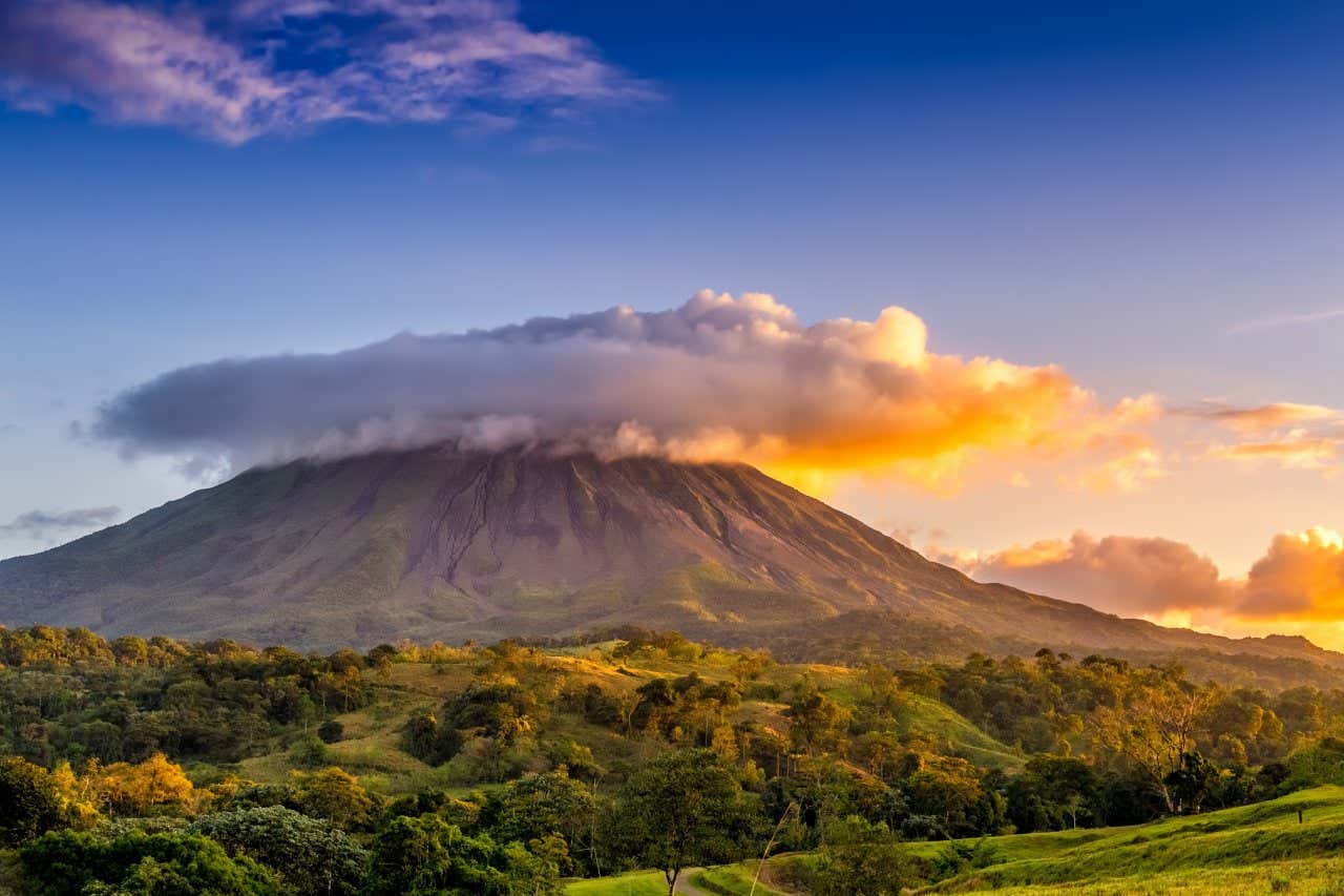 The Arenal Volcano as seen from afar, with clouds above it, against a mostly clear sky, and there is green vegetation in the foreground.