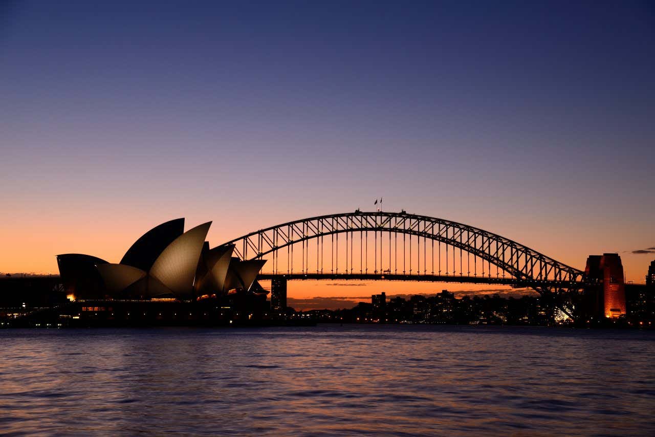 Sydney Harbour, with the Sydney Opera House and Harbour Bridge centred, seen at sunset, with an orange and purple sky in view.