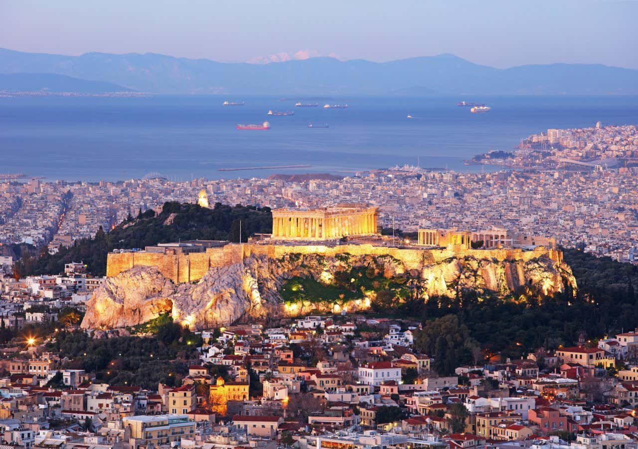 The Athens Acropolis lit up as night falls, as seen from afar, with the sea in view, with various ships sailing.