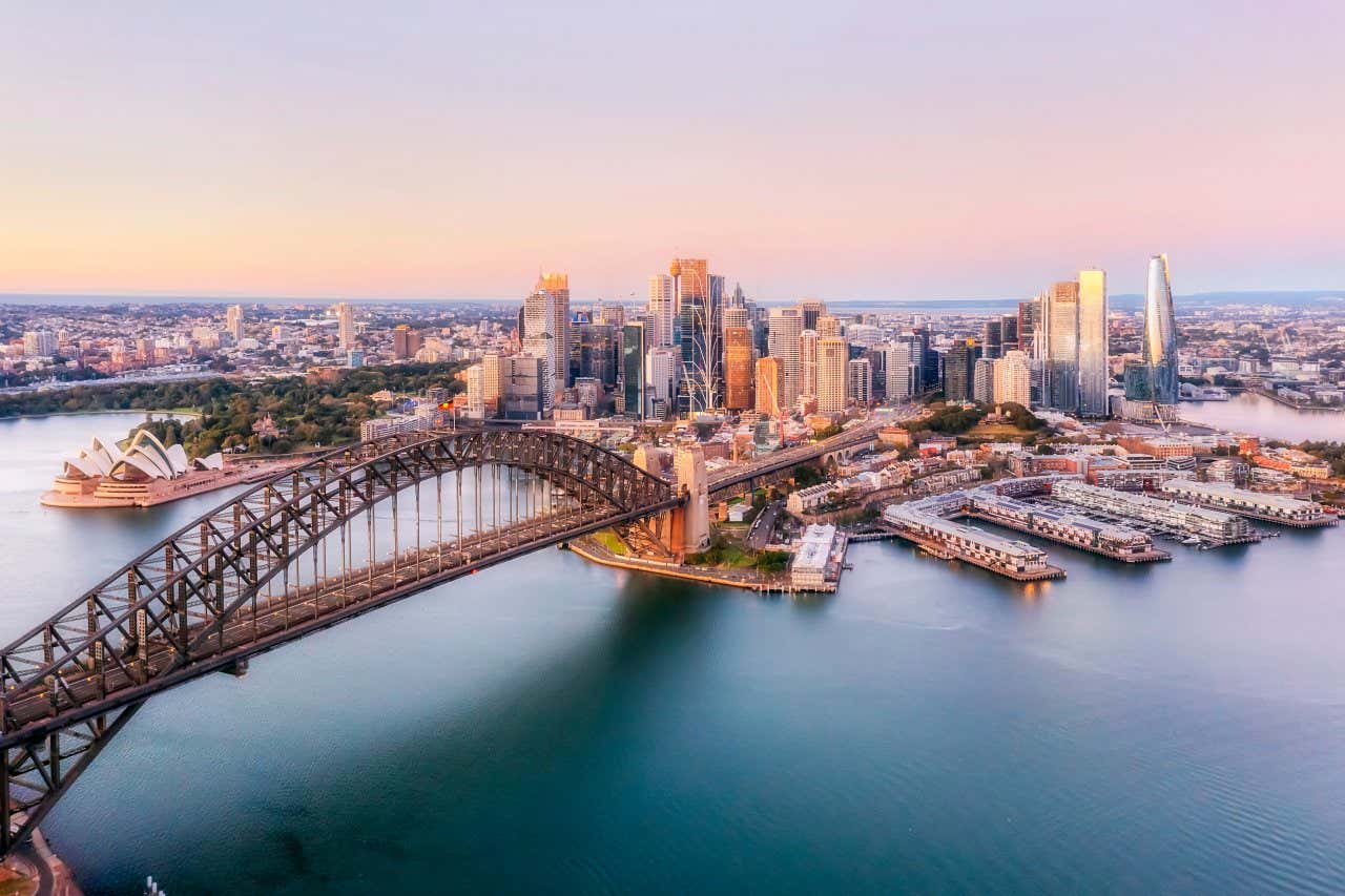 An aerial shot of Harbour Bridge, with Sydney Opera House and the city in the background, with a mostlypink sky.