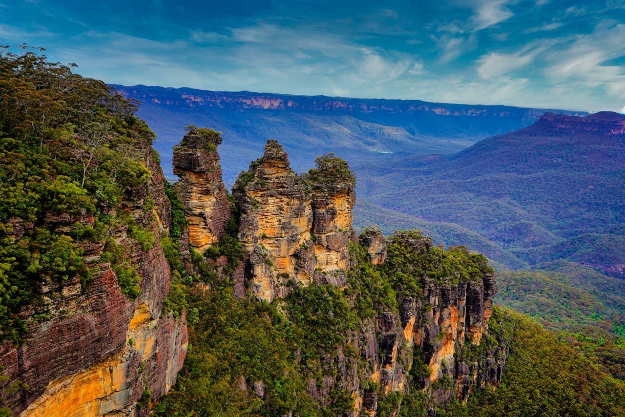 An aerial shot of the Three Sisters in Blue Mountains National Park, with mountainous landscape and a cloudy sky in the background.