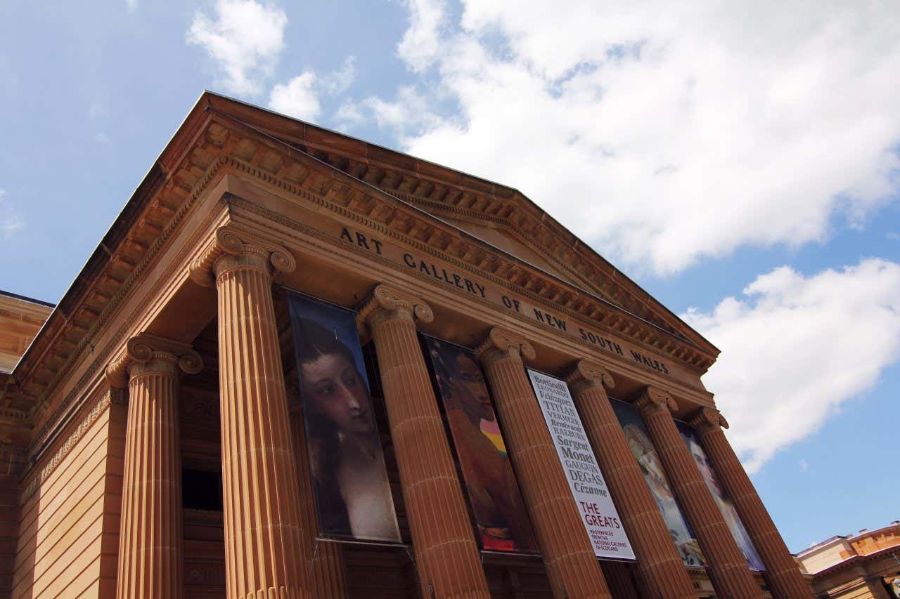 Art Gallery NSW, with ionic columns and some advertisements as seen from the ground, and a cloudy sky in the background.