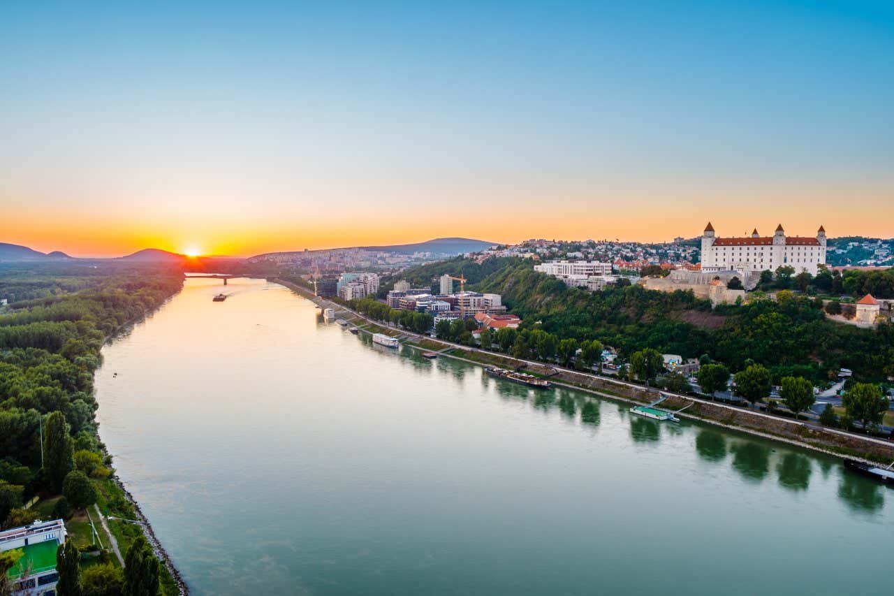 Danubio al tramonto circondato da alberi, con il castello di Bratislava sulla destra e una barca che solca il fiume in fondo, vicino al sole che tramonta