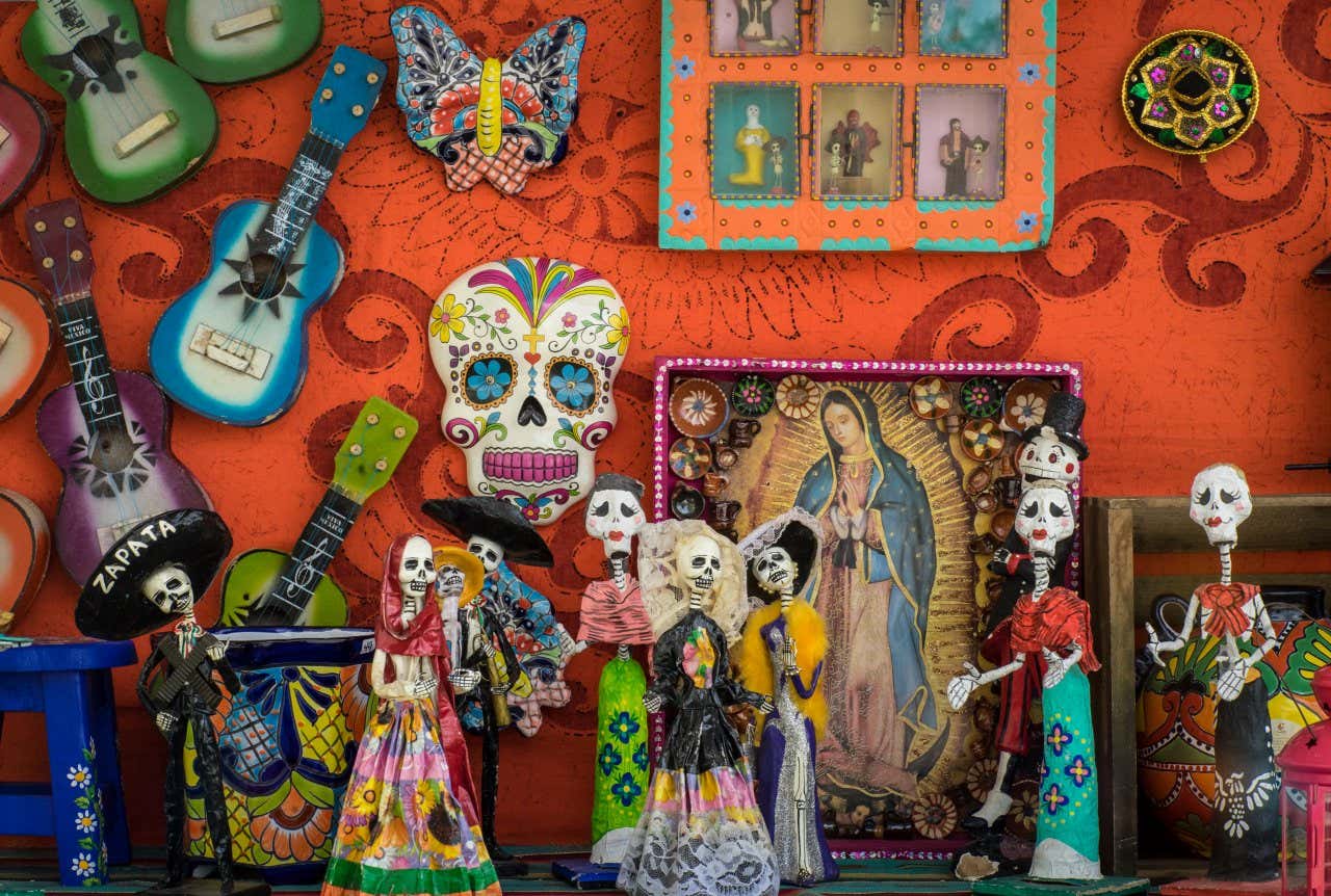 Multiple figures with the typical La Catrina skull design in front of a red wall with skulls and guitars on it as decoration.